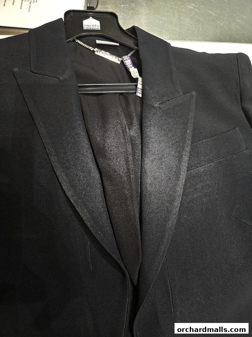 A close-up shot of a black suit jacket on a hanger with a tag from Presto Drycleaners.
