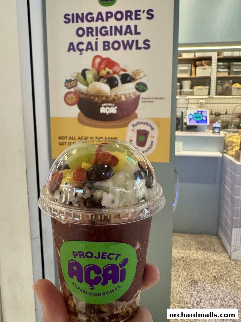 A hand holds a cup of acai bowl with fruits and granola, with a poster in the background.