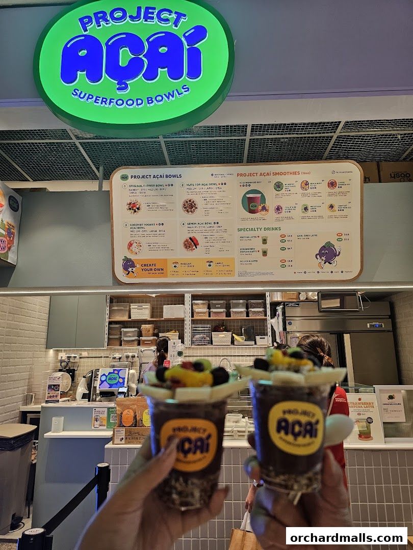 Two acai bowls are held up in front of the Project Acai shop counter and menu.
