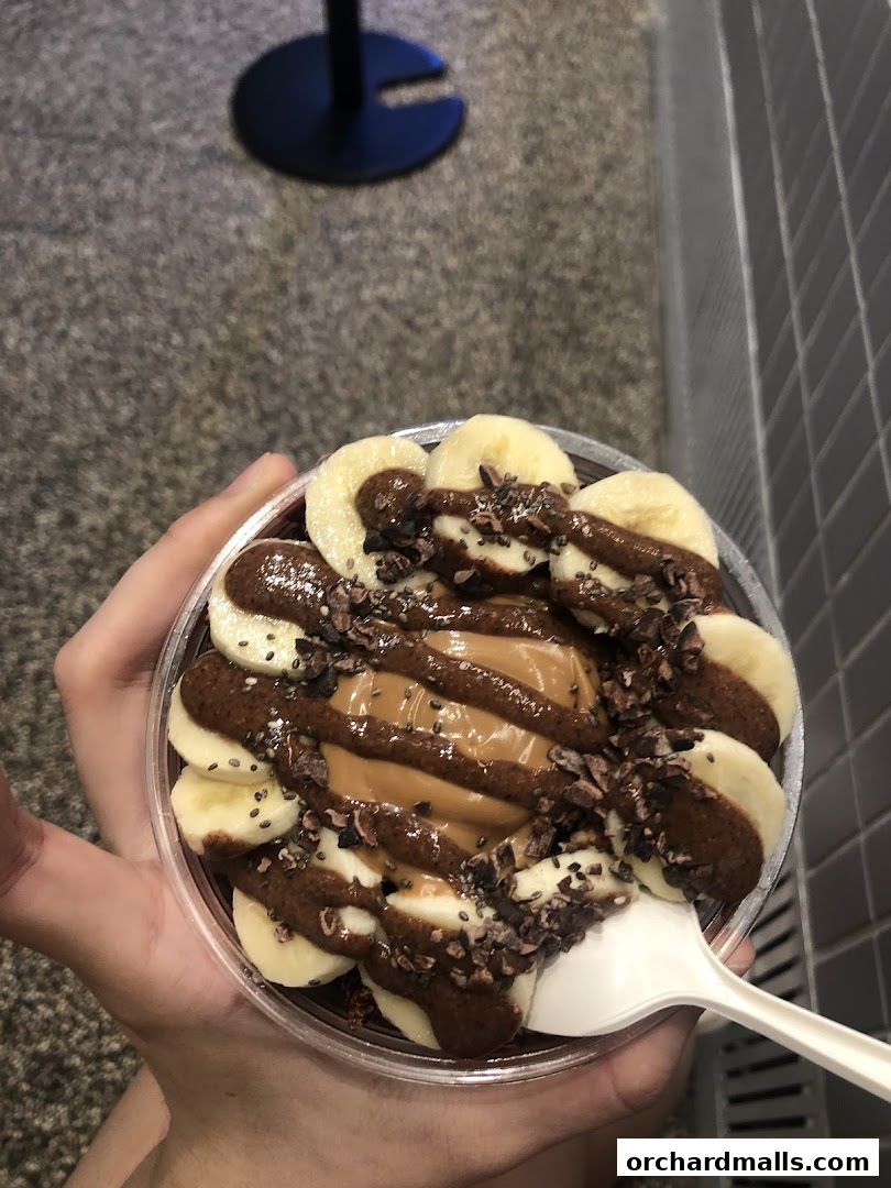 A hand holds a clear cup filled with acai, topped with banana slices, chocolate drizzle, and cacao nibs.