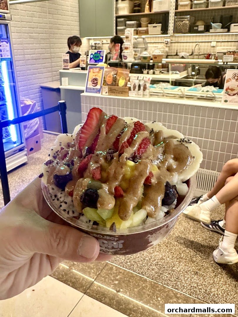 A hand holds a colorful acai bowl topped with fruits and sauce in front of a shop.