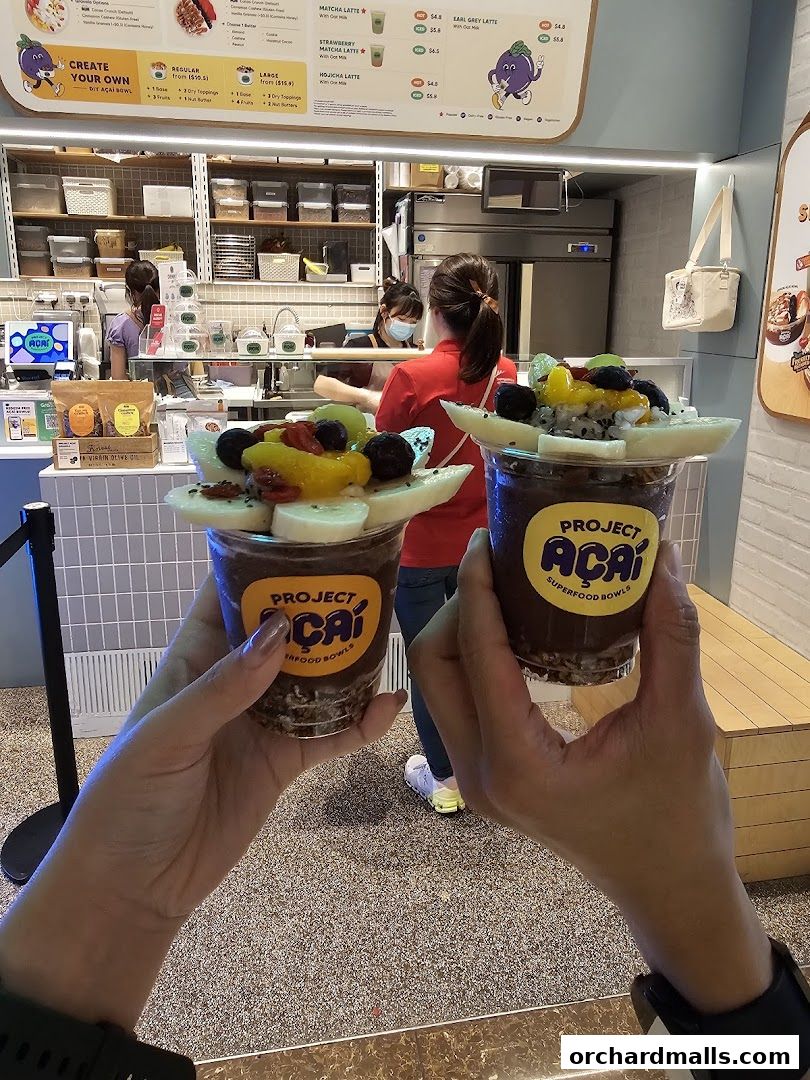 Hands holding two acai bowls with fresh fruit toppings in front of a shop counter.
