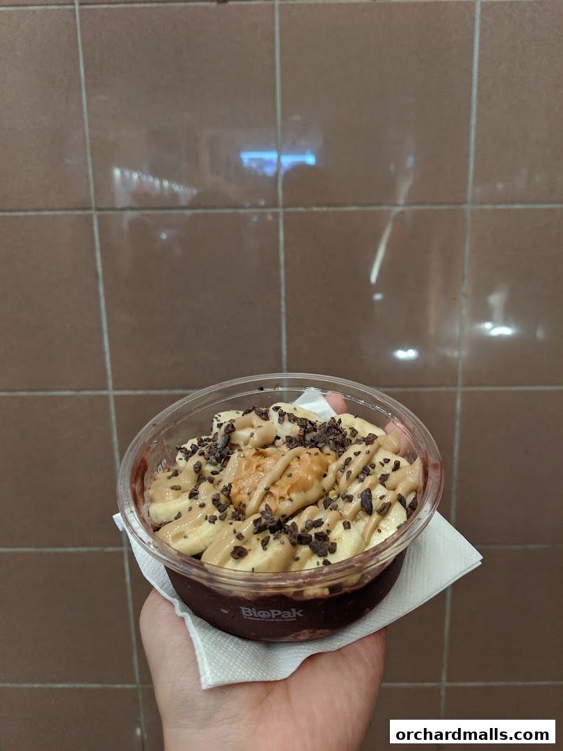 A hand holds a clear cup filled with acai, topped with banana slices, peanut butter, and cacao nibs.