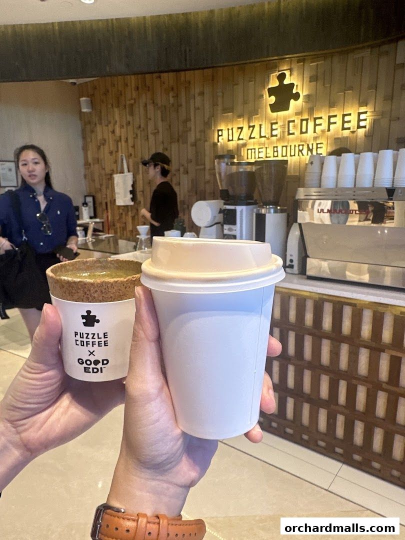 Two hands hold coffee cups in front of a coffee shop counter with a prominent sign.