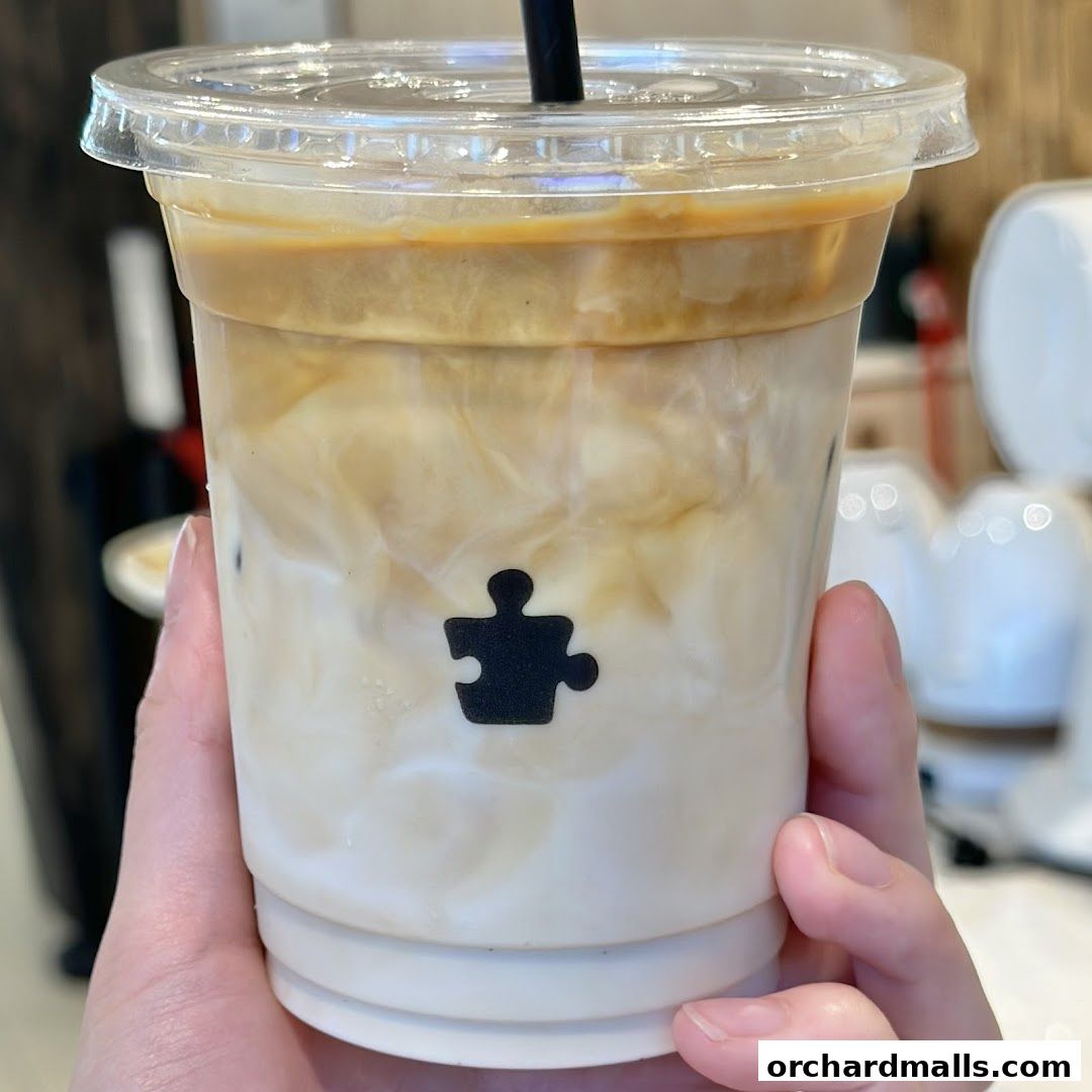 A hand holds a clear cup of iced coffee with milk and espresso layers, featuring a puzzle piece logo.