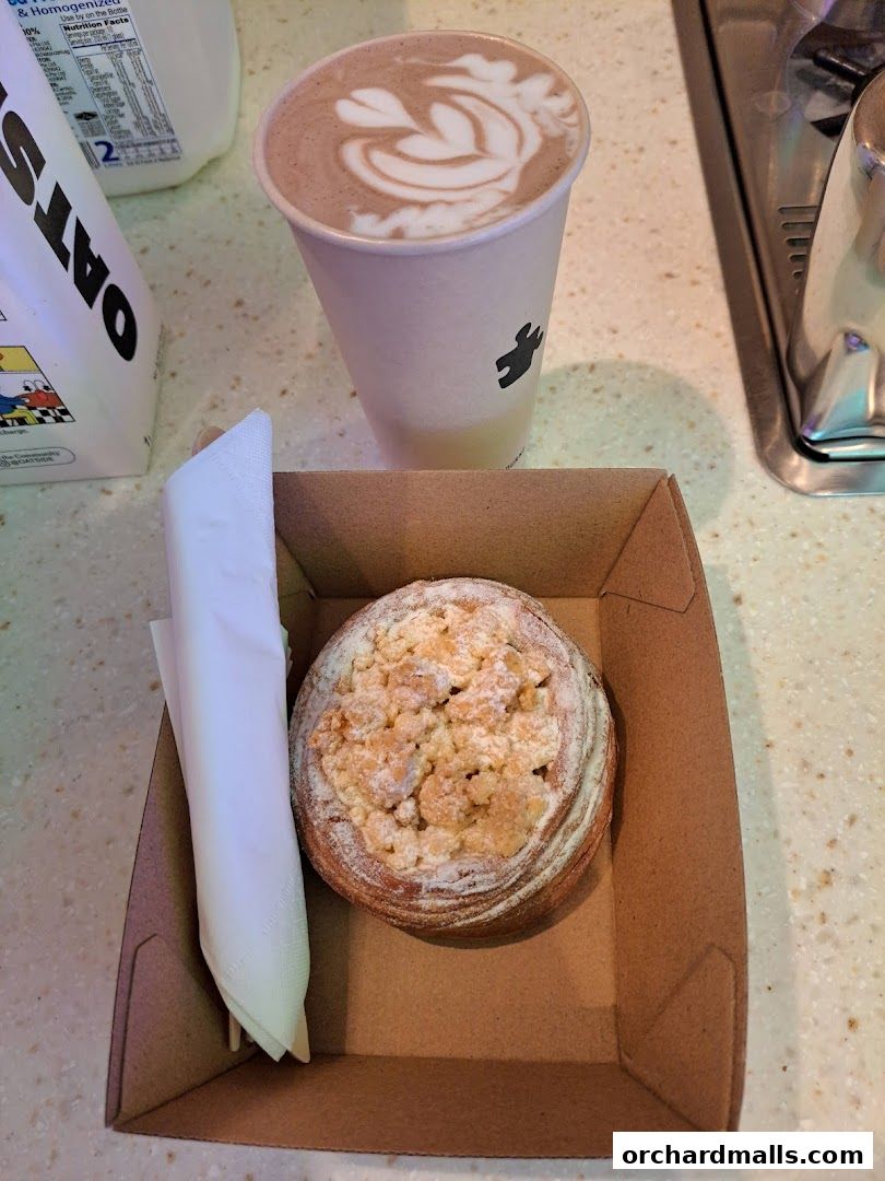 A coffee with latte art and a pastry in a box, next to a carton of oat milk.