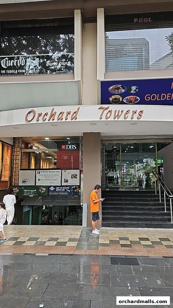 The entrance to Orchard Towers, featuring various shop signs and a man looking at his phone.