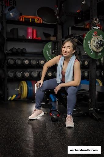 A smiling woman in workout clothes sits on a bench in a gym with weights and equipment.