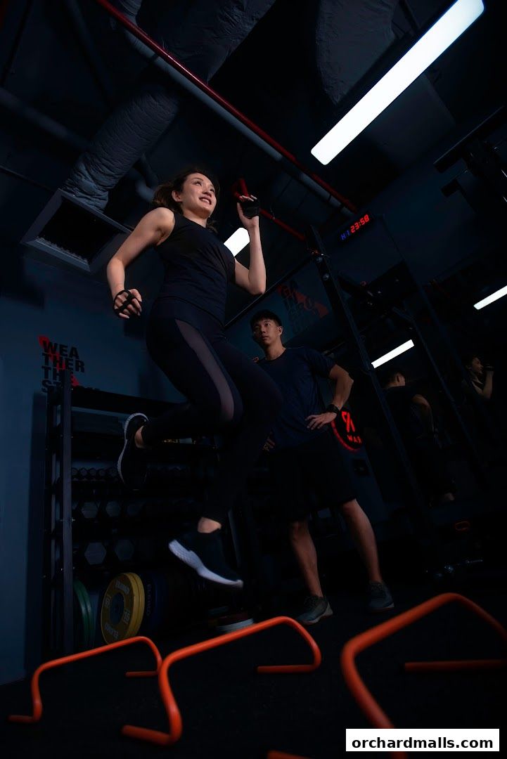 A woman jumps over hurdles while a man watches in a modern gym setting.