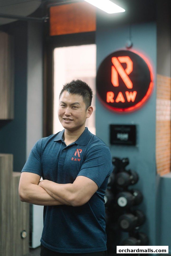 A man in a blue polo shirt with the RAW logo stands with his arms crossed in a gym.
