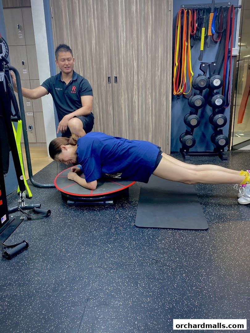 A personal trainer guides a client through a plank exercise on a balance board.
