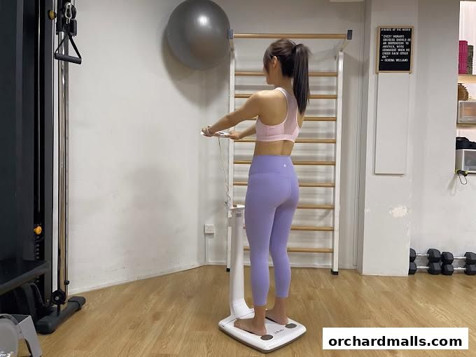 A woman stands on a body composition scale in a gym with exercise equipment.