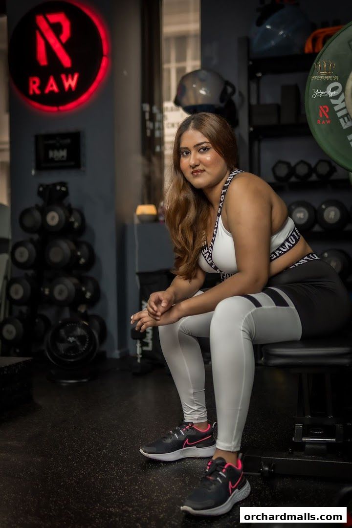 A woman in workout attire sits on a bench in a gym with weights in the background.