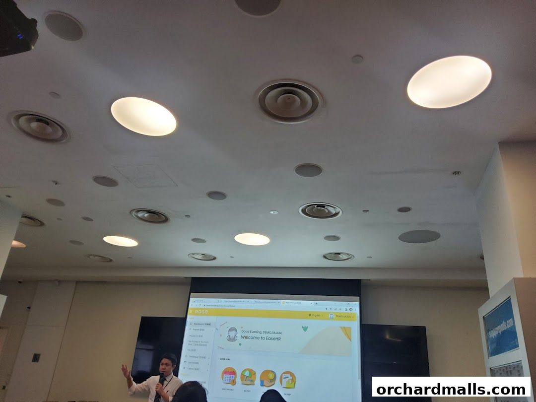 A man presents a digital interface on a projector screen in a room with many lights.
