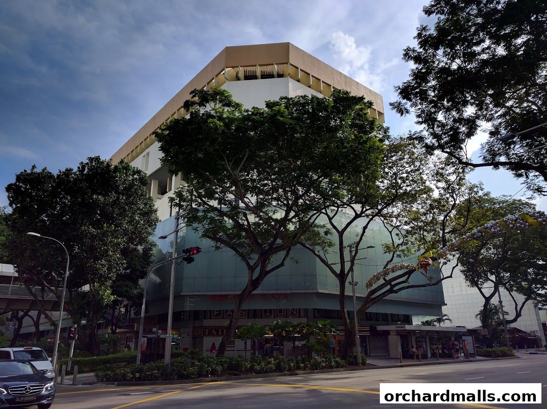 A modern building with glass facade and trees in front, under a cloudy sky.