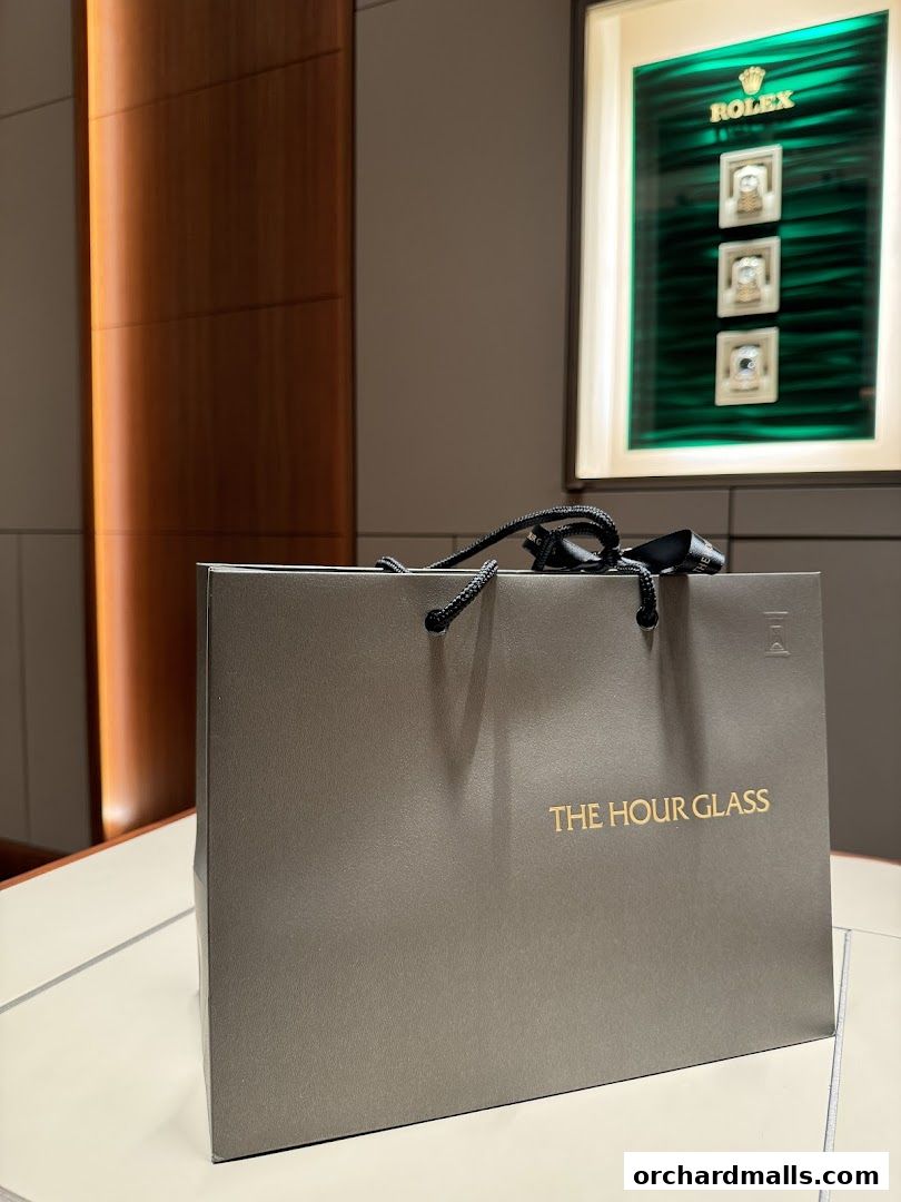 A shopping bag from The Hour Glass with a Rolex display in the background.