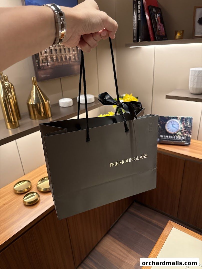 A person holds a shopping bag from The Hour Glass boutique, with luxury watches and decor in the background.