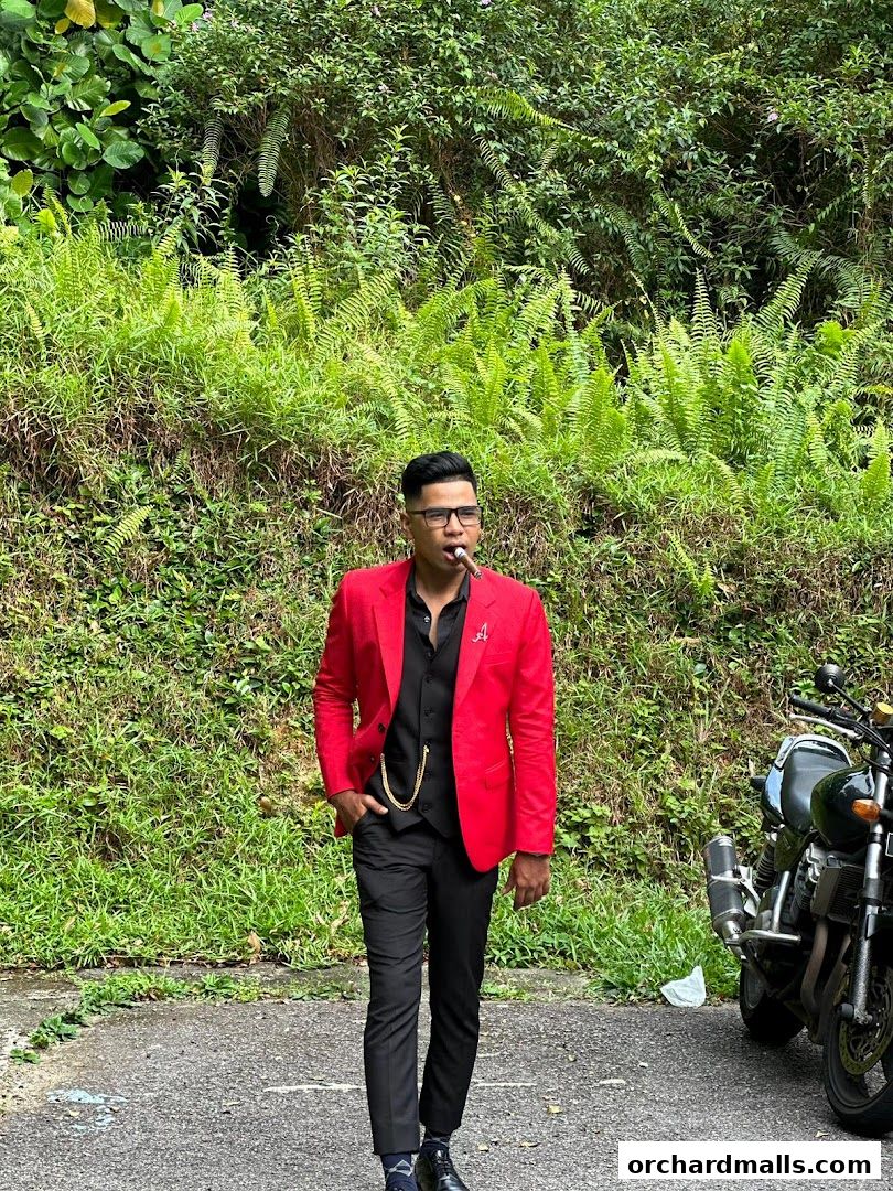 A man in a red suit and black vest smokes a cigar next to a motorcycle.