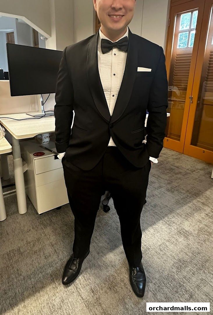 A man in a black tuxedo with a bow tie and white shirt stands with hands in pockets.