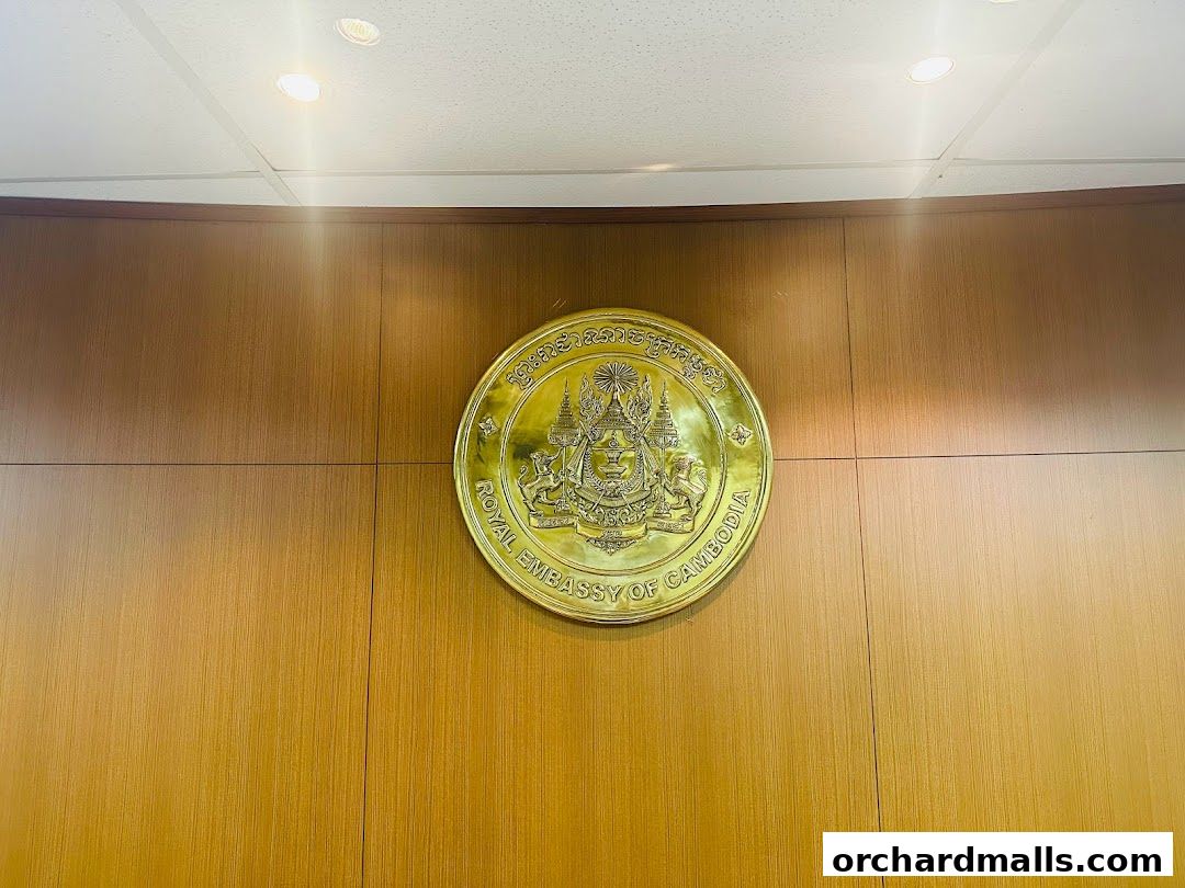 A golden emblem of the Royal Embassy of Cambodia mounted on a wood-paneled wall.