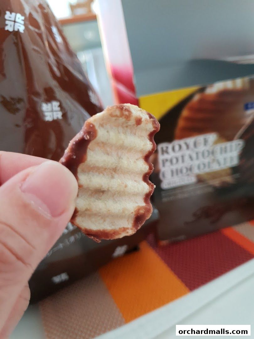 A hand holds a chocolate-covered potato chip with a box of Royce Potatochip Chocolate in the background.