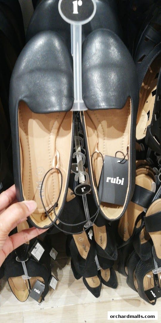 A hand holds a pair of black flat shoes from Rubi Shoes, displayed with other footwear.