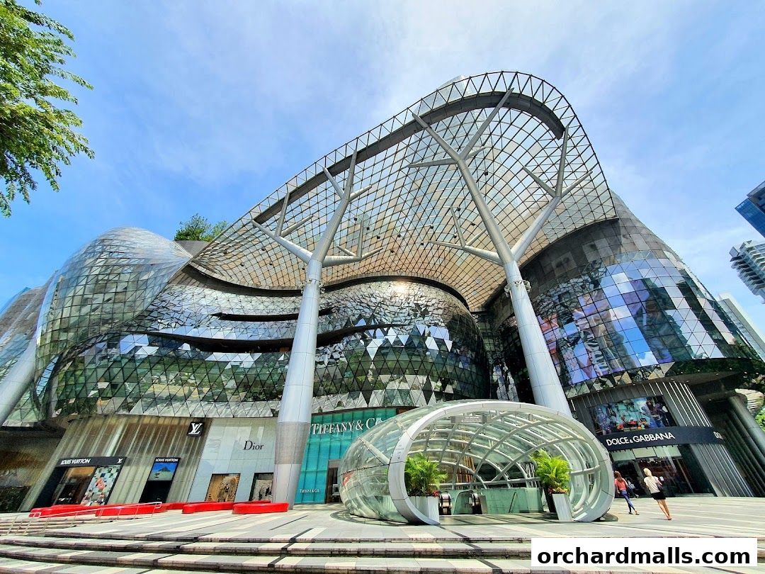 A modern shopping mall with a distinctive glass facade and multiple luxury brand storefronts.