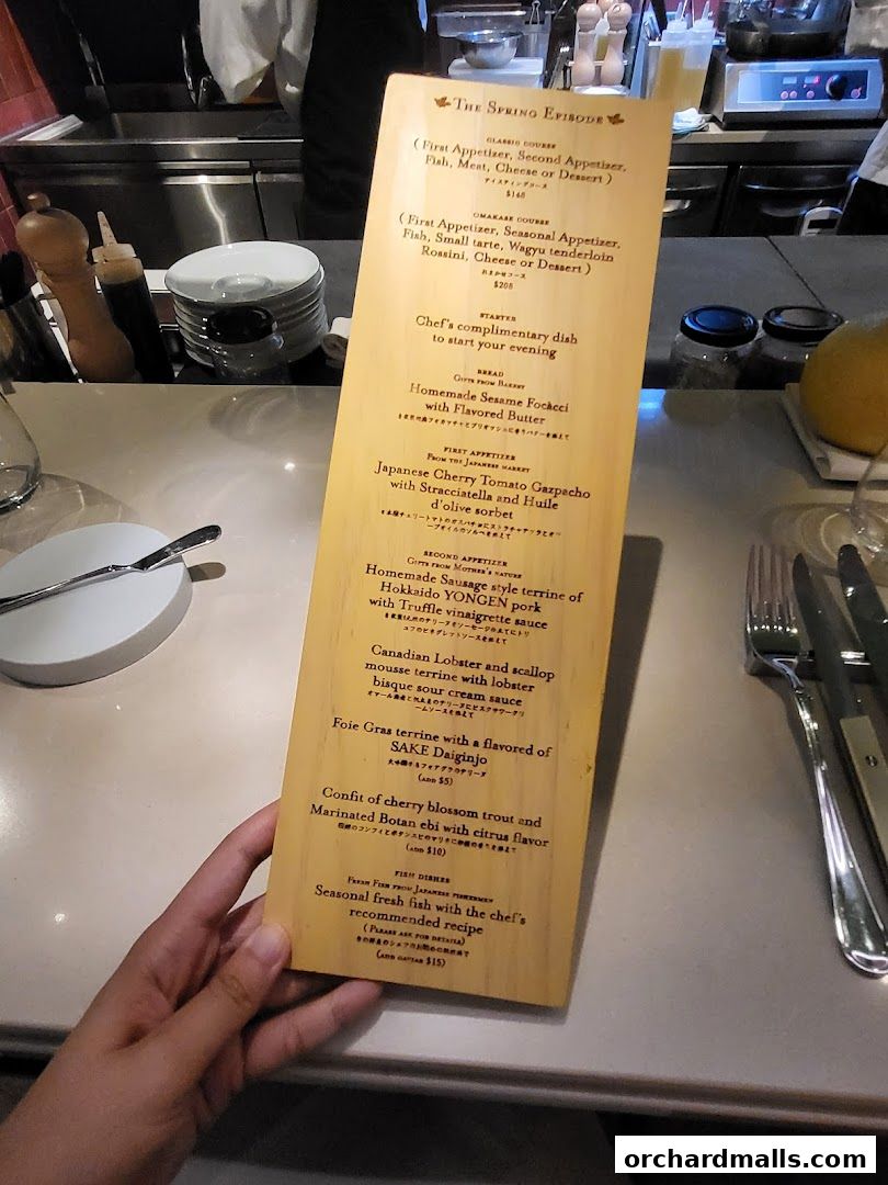 A hand holds a wooden menu titled 'The Spring Episode' with various dishes listed.