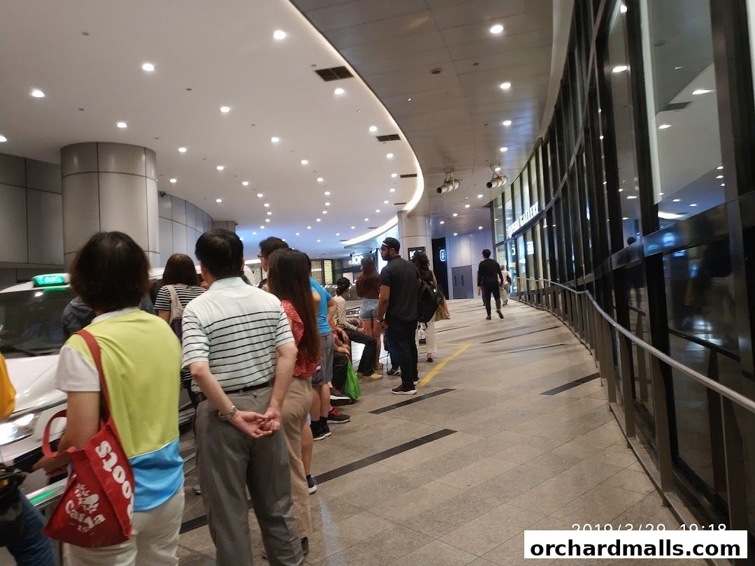 People queueing for a taxi in a modern shopping mall with a curved glass facade.