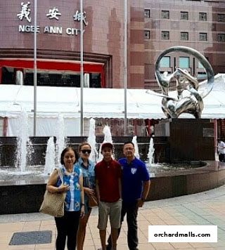 A family poses in front of the Ngee Ann City building with a fountain and sculpture.