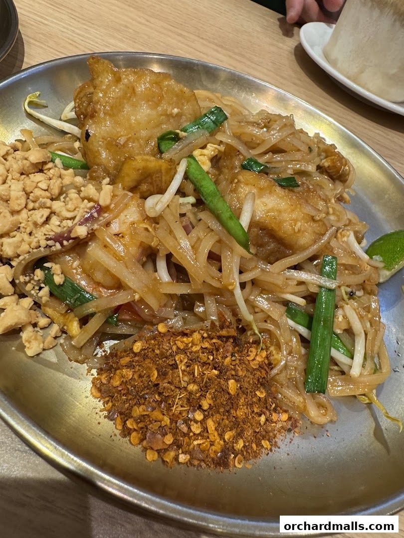 A close-up of Pad Thai with fried fish, peanuts, chili flakes, and lime.