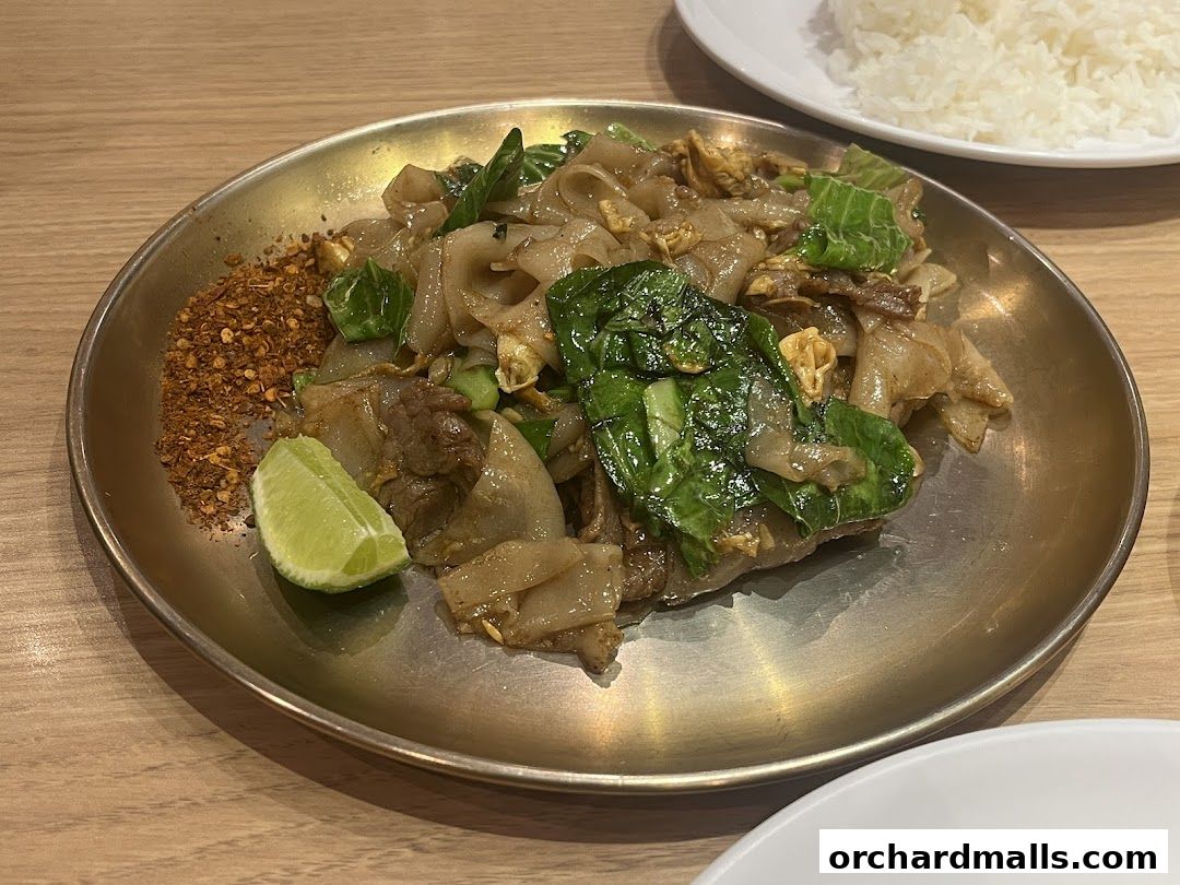 A plate of Pad See Ew with beef, Chinese broccoli, and chili flakes, served with a lime wedge.