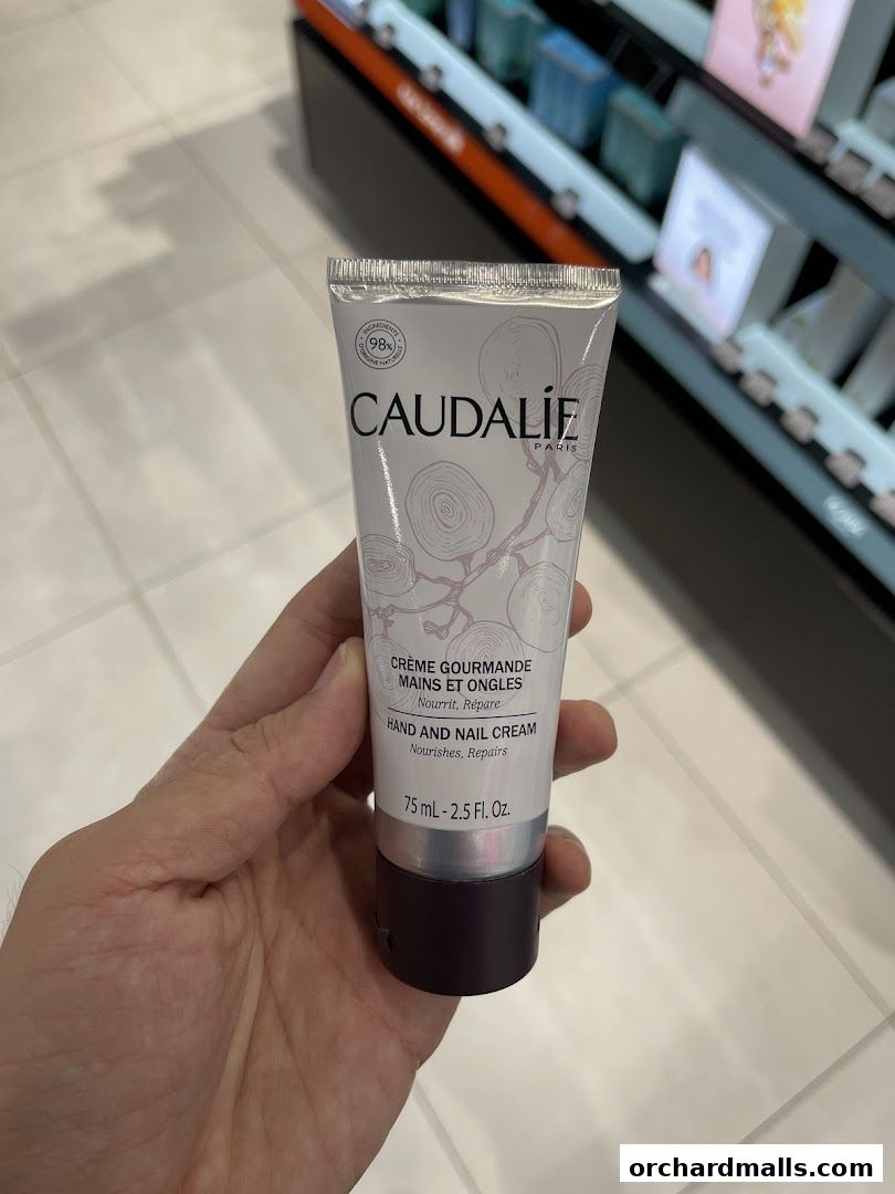 A hand holds a Caudalie hand and nail cream tube in front of a blurred retail display.