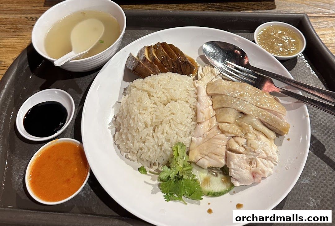 A plate of Hainanese chicken rice with soup and condiments on a tray.