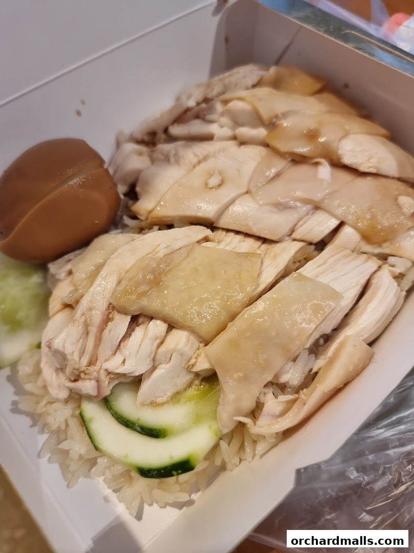 A close-up of a takeaway box filled with Hainanese chicken rice, featuring tender chicken slices, rice, cucumber, and a braised egg.