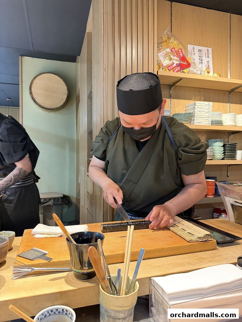 A sushi chef prepares a roll behind a counter with various tools and ingredients.