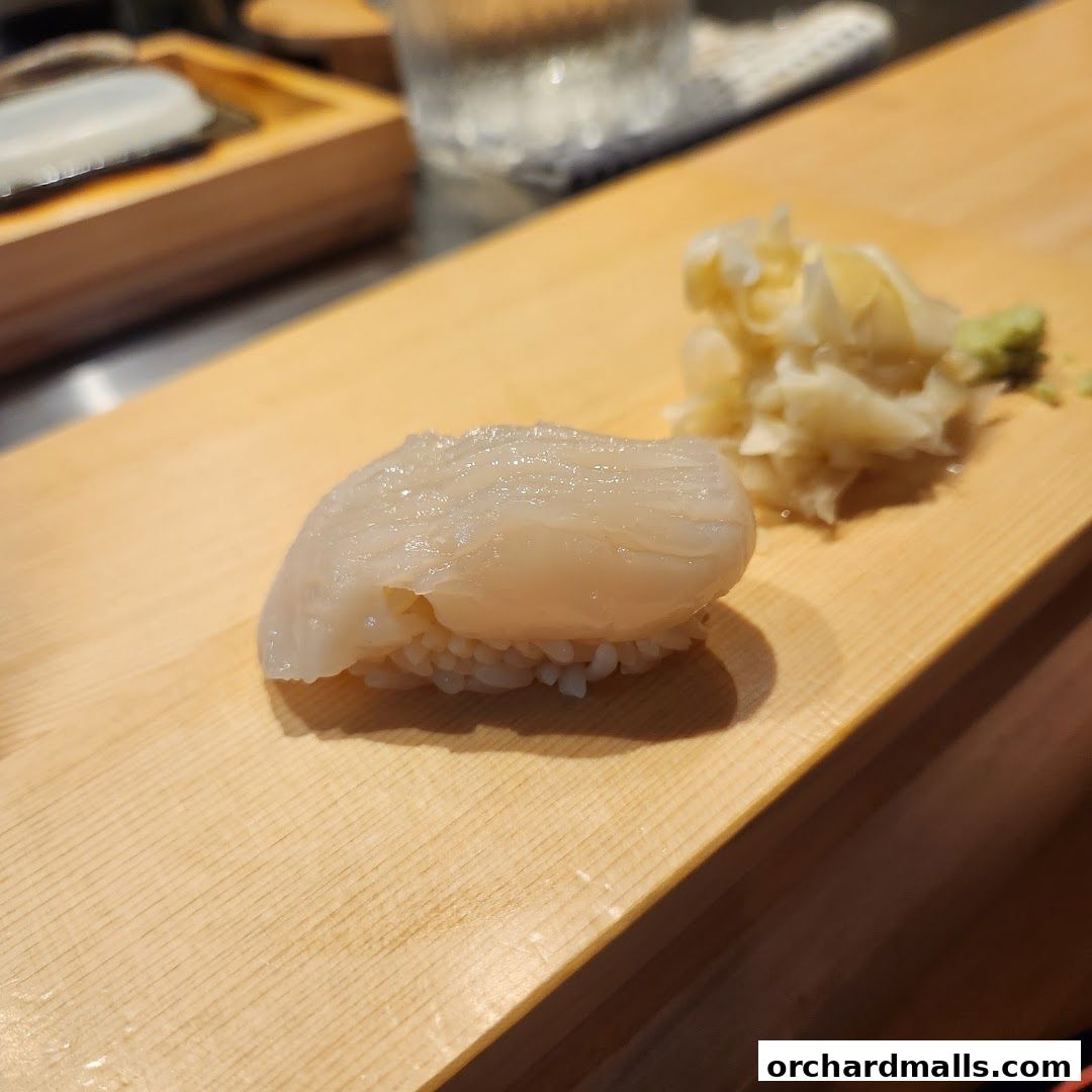 A close-up of a single piece of scallop sushi with ginger and wasabi on a wooden board.