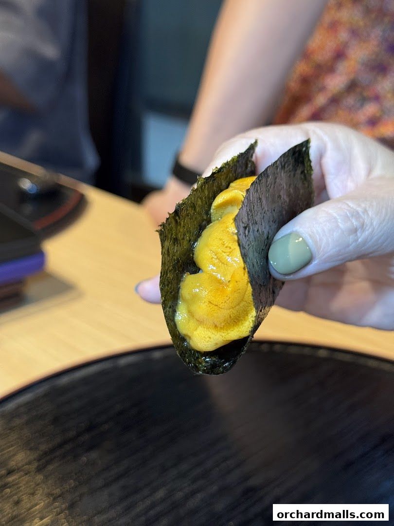 A hand holds a piece of sushi wrapped in nori, filled with bright orange uni.
