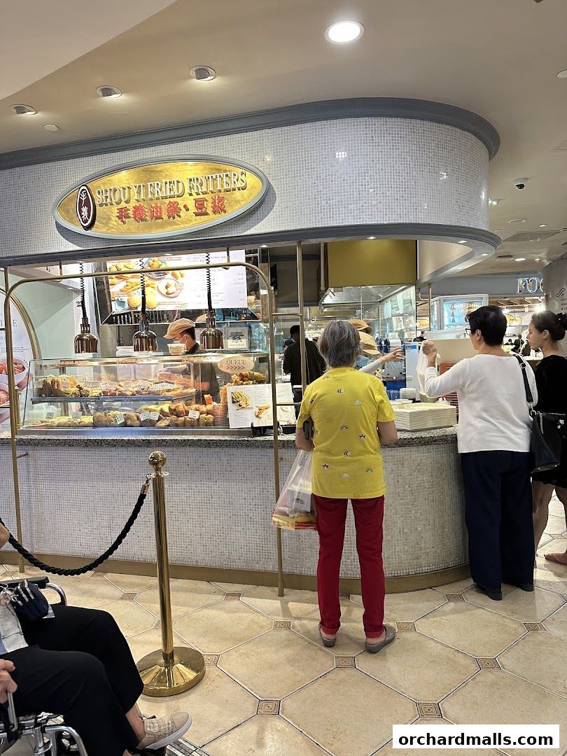 Customers queue at Shou Yi Fried Fritters, a shop displaying various fried snacks and drinks.