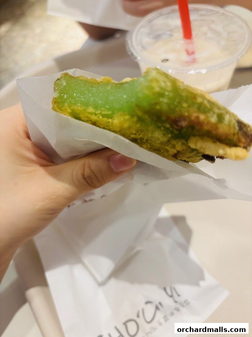 A hand holds a green fritter wrapped in white paper, with a drink in the background.