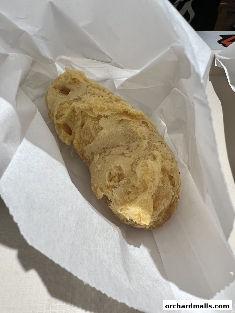 A close-up shot of a golden-brown, crispy fried fritter wrapped in white paper.