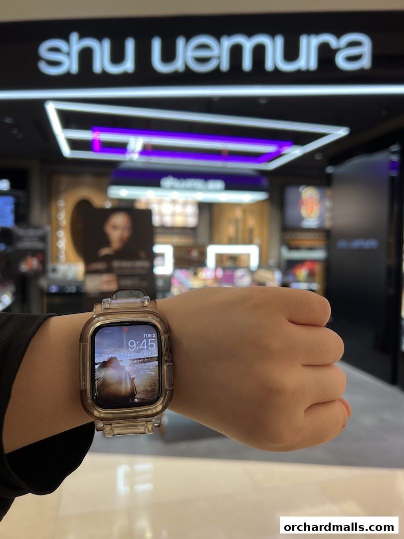 A person's wrist wearing a smartwatch in front of a Shu Uemura store.