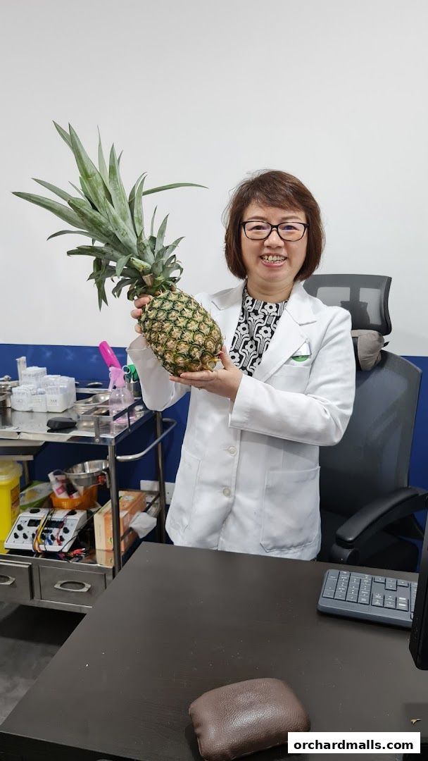 A smiling practitioner in a white coat holds a large pineapple in a clinic setting.