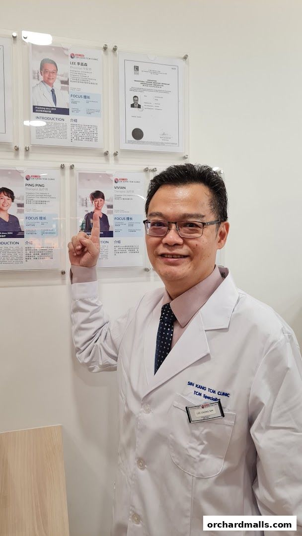 A TCM specialist in a white coat stands in front of framed credentials and staff profiles.