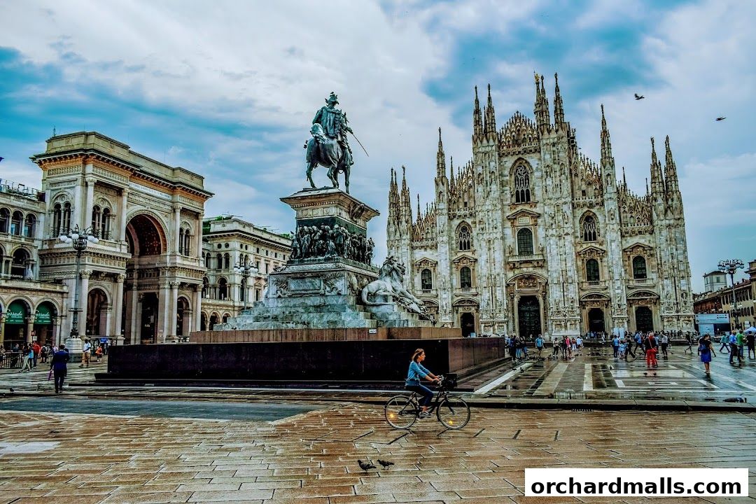 A person cycles past the Milan Cathedral and a monument in a public square.