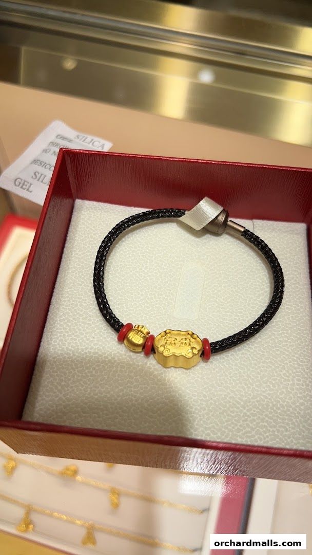 A close-up of a gold bracelet with a black braided band and red accents, presented in a red jewelry box.