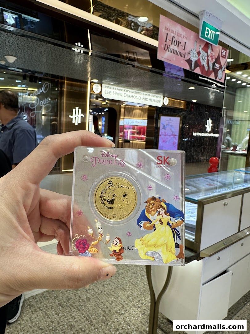 A hand holds a Disney Princess Belle and Beast gold coin in front of a jewelry store.