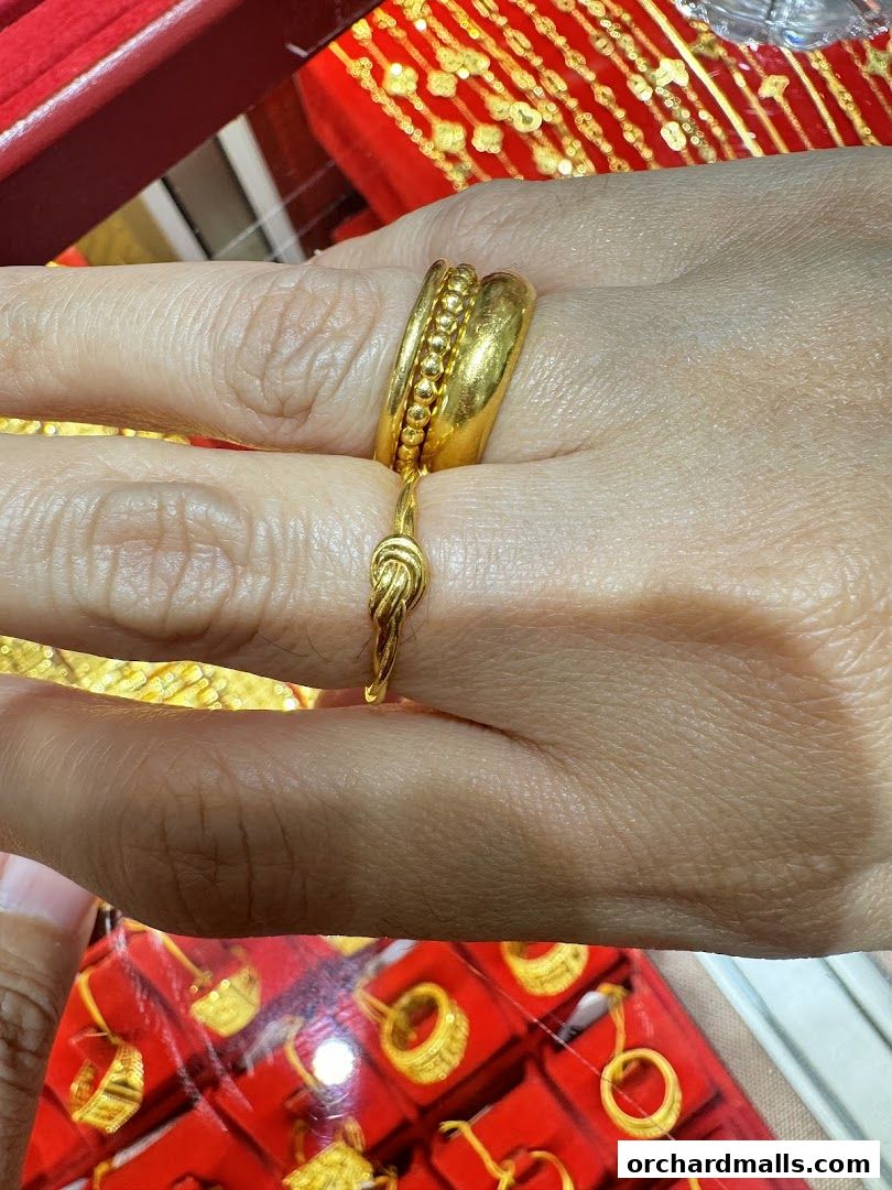A close-up of a hand wearing two gold rings, with more gold jewelry displayed in the background.