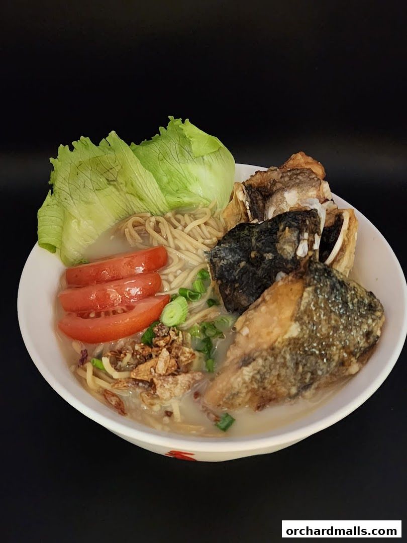 A bowl of fish soup with noodles, fried fish pieces, tomatoes, lettuce, and spring onions.