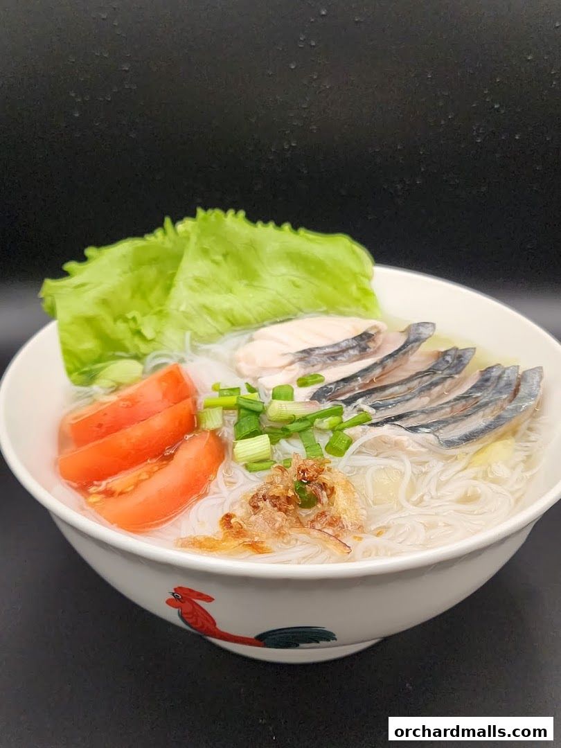 A bowl of fish soup with noodles, fish slices, tomatoes, lettuce, and spring onions.